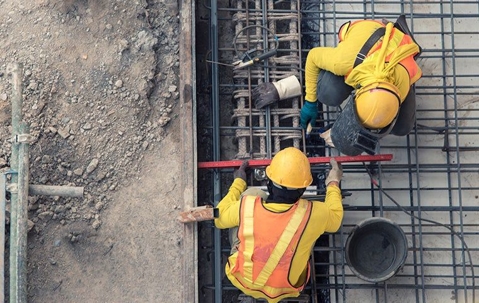 Construction workers wearing bright yellow shirts, safety helmets, and reflective vests while welding metal framing on the ground.